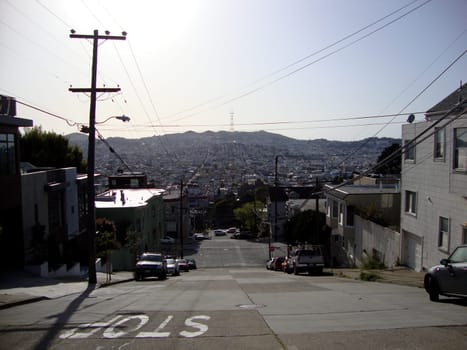 San Francisco - March 26, 2010:  A bright and cheerful photo of a street in San Francisco, California. The street is sloping downwards and has a panoramic view of the city skyline in the background. The street is lined with white houses and cars parked on the side. The houses have a modern and elegant design. The street has power lines running above it, adding a touch of urban charm. The sky is blue and the sun is shining, creating a contrast between the light and the shadow. 