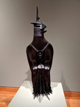 Seattle - May 16, 2019:  War God, a sculpture by Philip McCracken, displayed at the Seattle Art Museum as part of the exhibition "American Art: The Stories We Carry". The sculpture is a carved figure in cedar wood with a leather strap and saw blades as accessories, representing the forces of "anti-life". The sculpture has a dark, significant presence, contrasted by the white wall and the wooden floor of the gallery. The sculpture is lit from above, creating a shadow on the wall behind it.