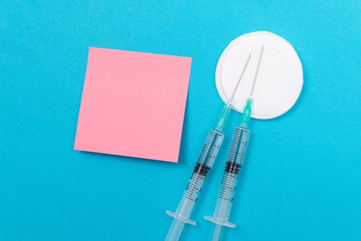 Vaccination, Immunology or Revaccination Concept - Two Medical Disposable Syringe Lying on Blue Table in Doctor's Office in a Hospital or Clinic. Blank Pink Sticky Note - Mock Up with Copy Space