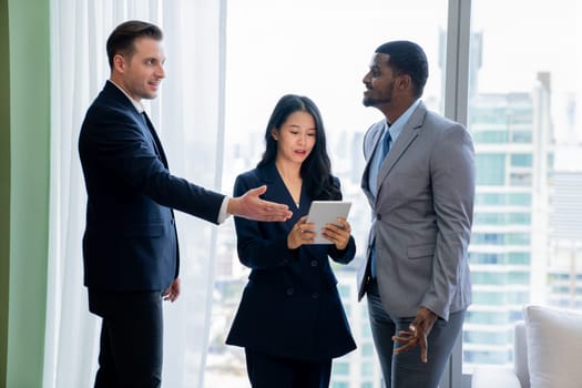 Diverse smart businesspeople standing near window with skyscraper while looking at business data analysis. Group of business team using tablet represent marketing plan, strategy, idea. Ornamented..