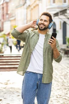 Happy relaxed overjoyed Caucasian young man in wireless headphones choosing, listening favorite energetic disco rock n roll music dancing outdoor. Guy standing in urban city sunshine street. Vertical