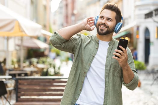 Happy relaxed overjoyed Caucasian man in wireless headphones choosing, listening favorite energetic disco music in smartphone dancing outdoors. Guy tourist walking passes by urban sunshine city street
