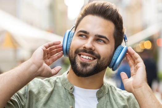 Happy relaxed overjoyed Caucasian young man in wireless headphones choosing, listening favorite energetic disco rock n roll music dancing outdoor. Guy standing in urban city sunshine street background