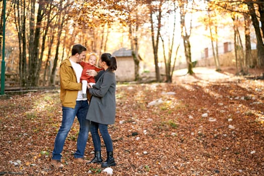 Mom and dad are holding a little girl in their arms while standing with her on fallen leaves in the park. High quality photo