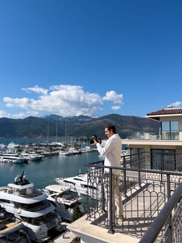Young man stands on a balcony and takes pictures of luxury yachts moored off the coast. High quality photo