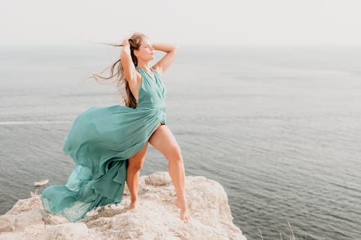 Side view a Young beautiful sensual woman in a mint long dress posing on a volcanic rock high above the sea during sunset. Girl on the nature on overcast sky background. Fashion photo