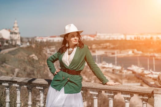 Woman walks around the city, lifestyle. Happy woman in a green jacket, white skirt and hat is sitting on a white fence with balusters overlooking the sea bay and the city