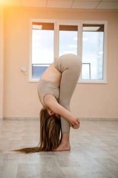 Young woman doing yoga in the gym. A girl with a long braid and in a beige tracksuit stands in a stork pose on a pink carpet. A woman performs padahstasana