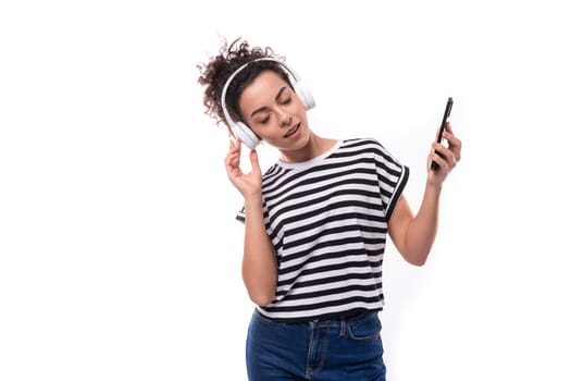 young well-groomed curly woman with black hair in a summer t-shirt listens to music in wireless headphones and holds a phone.