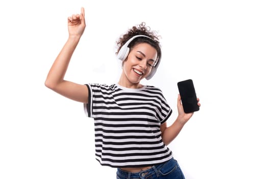 young caucasian woman with curly haircut dressed in a striped summer t-shirt with wireless headphones shows ads on a smartphone screen with a mockup.