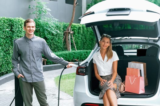 Young couple travel with EV electric car charging in green sustainable city outdoor garden in summer shows urban sustainability lifestyle by green clean rechargeable energy of electric vehicle innards