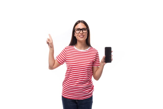 young charming brunette woman with glasses dressed in a striped t-shirt demonstrates the screen of a mobile phone on a white background with copy space.