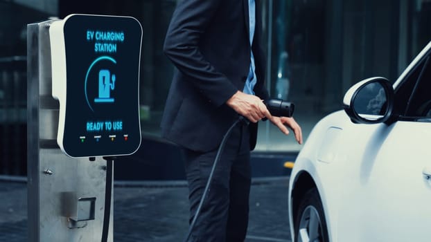 Businessman recharge his electric car from charging station at city center or public car park. Eco friendly rechargeable car using alternative clean energy in urban city lifestyle.Peruse