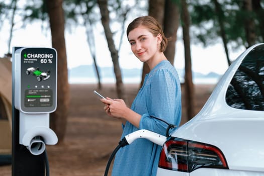 Holiday road trip vacation traveling to the beach camp with electric car, young woman checking battery from smartphone while recharge EV vehicle. Beach travel camping with eco-friendly car .Perpetual