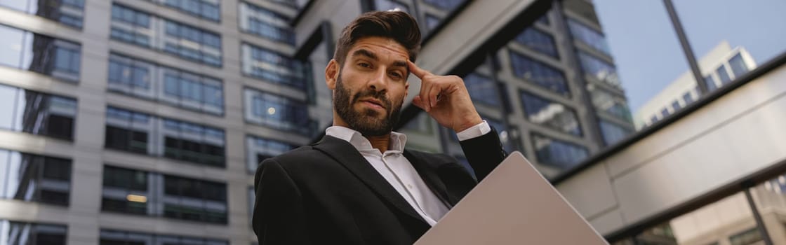 Handsome stylish businessman standing with laptop on office terrace and looks camera