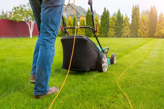A gardener with a lawn mower is cutting green grass in the garden, in the house backyard on a sunny summer day. The legs of the worker and a lawn mower, a close-up.