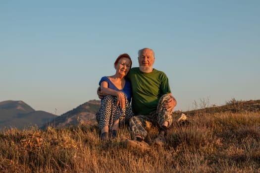 An elderly couple sits on a mountain with their backs with a beautiful view of the mountains in the distance