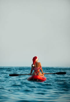 Sexy girl in a Santa Claus hat floats on the sea on a Sup Board in a red bikini celebrating Christmas at a resort in a hot country, Standup paddleboarding
