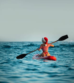 Sexy girl in a Santa Claus hat floats on the sea on a Sup Board in a red bikini celebrating Christmas at a resort in a hot country, Standup paddleboarding