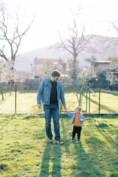 Dad with a little girl walks through the green pasture from the fence, holding hands. High quality photo