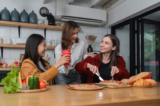 A group of young female friends have a party with pizza on the table and red drink glasses. Talk and live together happy, having fun at home..