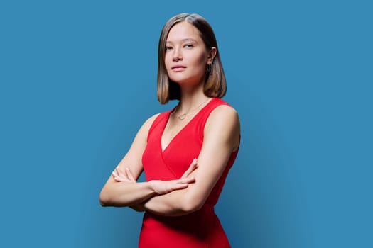 Young confident serious woman in red on blue studio background. Successful fashionable female with crossed arms looking at camera. Business, work, services, education, fashion beauty professions