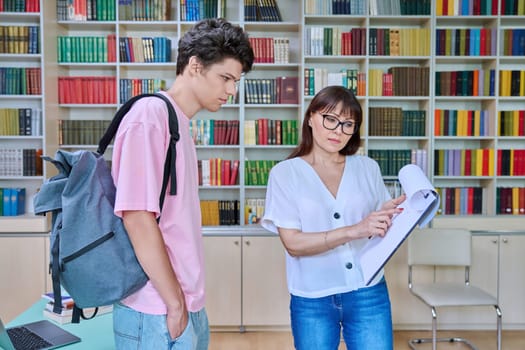 Middle-aged female teacher mentor talking to male college student, young 18-20 year old guy, inside library. Knowledge, education, youth, college university concept