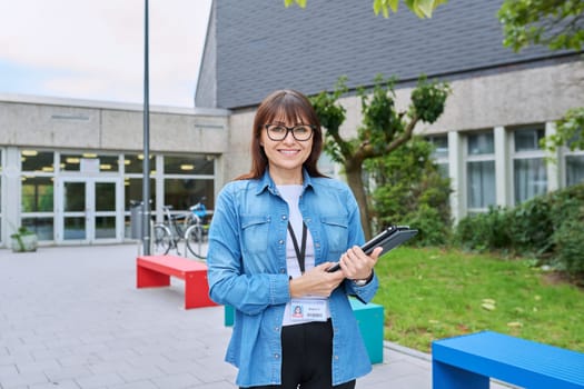 Middle-aged confident woman school teacher, mentor, pedagogue, psychologist, counselor, social worker with digital tablet in hands posing near school building, outdoor