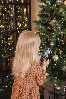 A little blonde girl photographs balloons on a Christmas tree in a festive interior decorated in a New Year's style. The concept of a merry Christmas