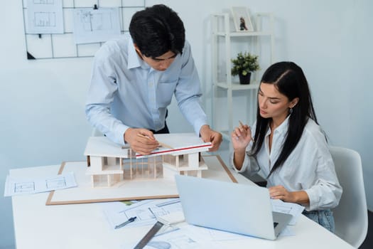 Professional male asian architect using ruler to measure house model length while young beautiful caucasian colleague using laptop to analyzed data on meeting table with house model. Immaculate.