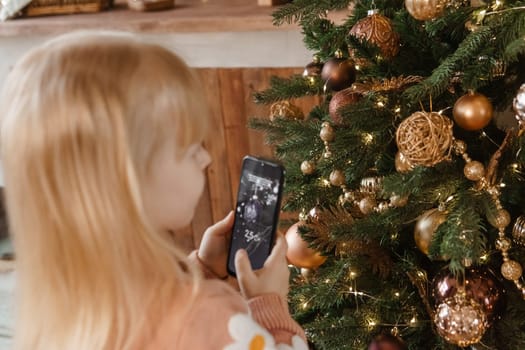 A little blonde girl photographs balloons on a Christmas tree in a festive interior decorated in a New Year's style. The concept of a merry Christmas