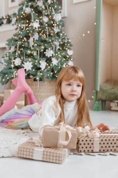 A little red-haired girl lies next to Christmas gifts in a craft package. Happy Christmas concept