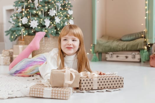 A little red-haired girl lies next to Christmas gifts in a craft package. Happy Christmas concept