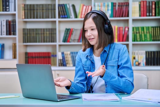 Young female college student in headphones sitting at desk with laptop having online video conference chat, looking at screen talking listening watching learning class, webinar, lesson inside library
