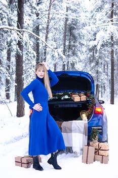 Woman in winter snowy forest in blue dress next to blue car decorated with Christmas decor. Christmas and winter holidays concept