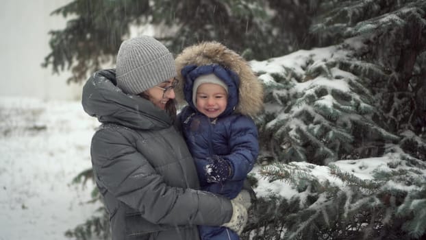 A young mother with her little son in her arms against a background of fir trees in winter. A woman and a child stand under the snow and laugh. 4k