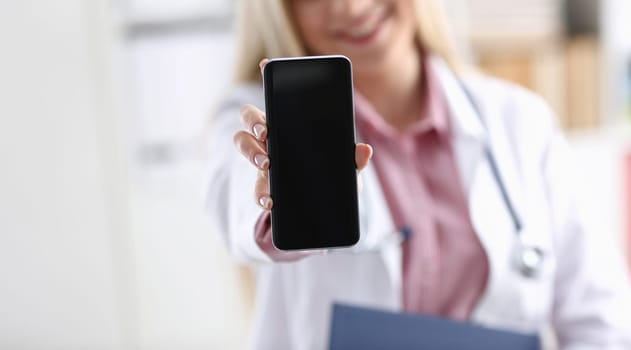 Beautiful female blonde doctor holding smartphone in hand at office woman discussing disease and giving an online consultation to a remote training mobile advertising app