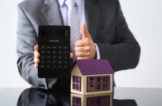 Man in suit and tie hold in arms calculator with little toy house closeup. New owner or tenant percent rate, sell or rental structure seller deal family solution pledge negotiation, industry