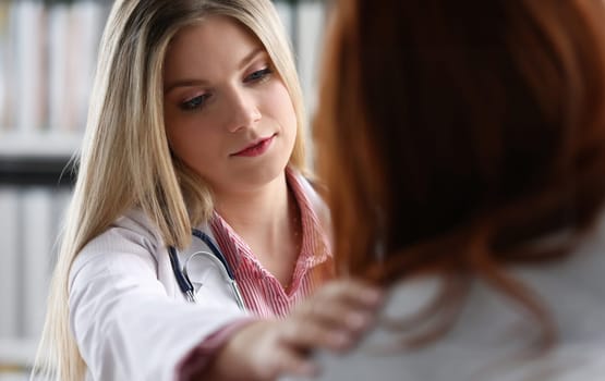 Handsome friendly female doctor talk with thankful female patient at office and greet her with hand on shoulder gesture. Welcoming friend trust physical healthy lifestyle exam interview concept