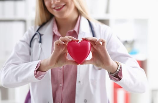 Beautiful smiling blond female doctor hold in arms red toy heart closeup. Cardio therapeutist student education CPR 911 life save physician make cardiac physical pulse rate measure arrhythmia