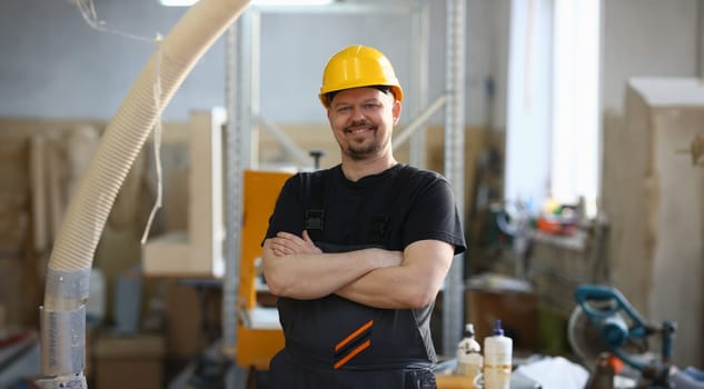Portrait of young attractive man in work clothes and yellow helmet crossed arms smiling at shop for manufacturing furniture and details home interior