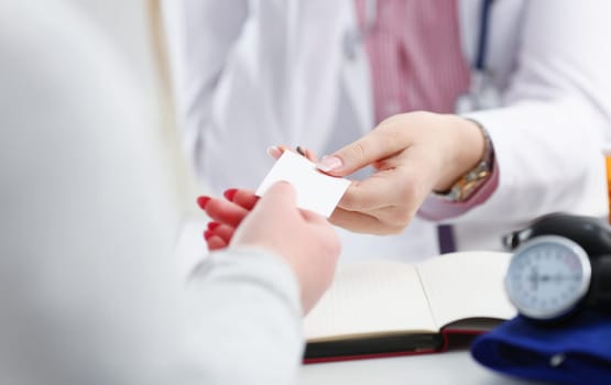 Female physician hand give white blank calling card to businesswoman closeup in office. Physical disease prevention examine patient instrument shop healthy lifestyle family doctor concept