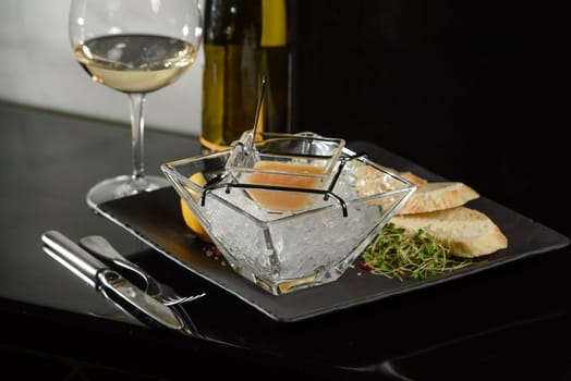 Pike fish caviar, on ice, with croutons and butter, on a transparent dish with a glass of white wine on a dark background