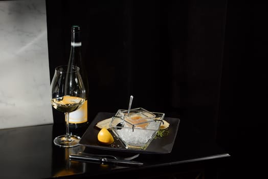 Pike fish caviar, on ice, with croutons and butter, on a transparent dish with a glass of white wine on a dark background