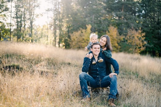 Smiling mom hugging dad from behind with little girl on shoulders sitting on stone on lawn. High quality photo