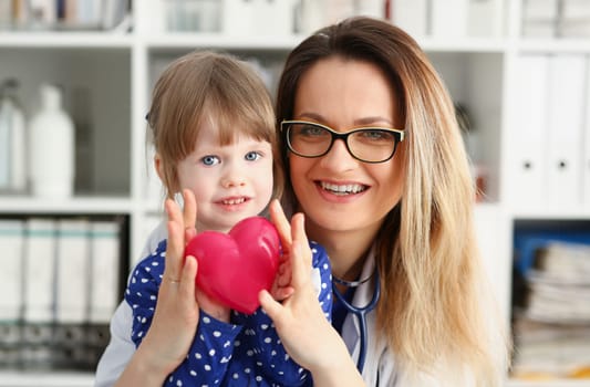 Female doctor and happy little child hold in arms red toy heart closeup. Cardio therapeutist student education CPR 911 life save physician make cardiac physical pulse rate measure arrhythmia lifestyle