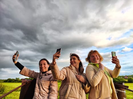 Three funny tourist girls on old bridge take selfies against the background of an alarming dramatic sky with thunderclouds. Middle-aged women having fun outdoors