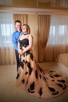 A plump woman in royal dress and a skinny man together in honeymoon hotel room. An adult newlywed couple embraces in an intimate setting. Queen Girl and guy in love