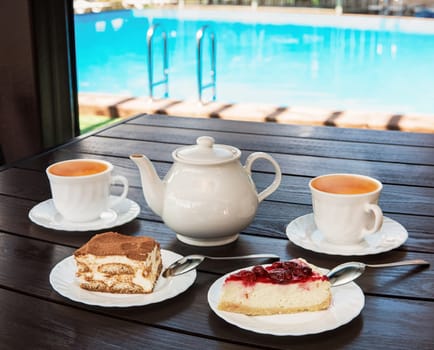 Afternoon tea set with hot tea and tasty cakes on the table and the swimming pool. Hotel resort or resting concept