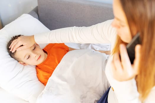 Worried young mother of sick child calling to a doctor on phone. Sick kid feels bad and lying in bed under a blanket at home.